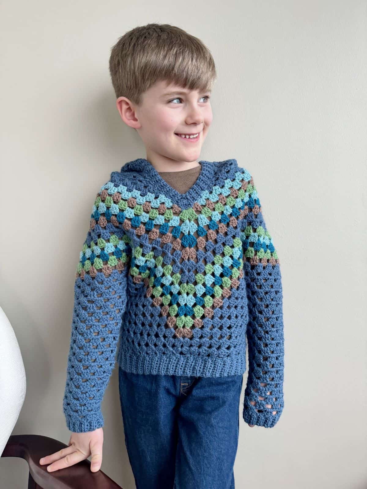 A young boy standing indoors wearing a blue, green, and brown crocheted sweater and blue jeans, smiling and looking to the side.