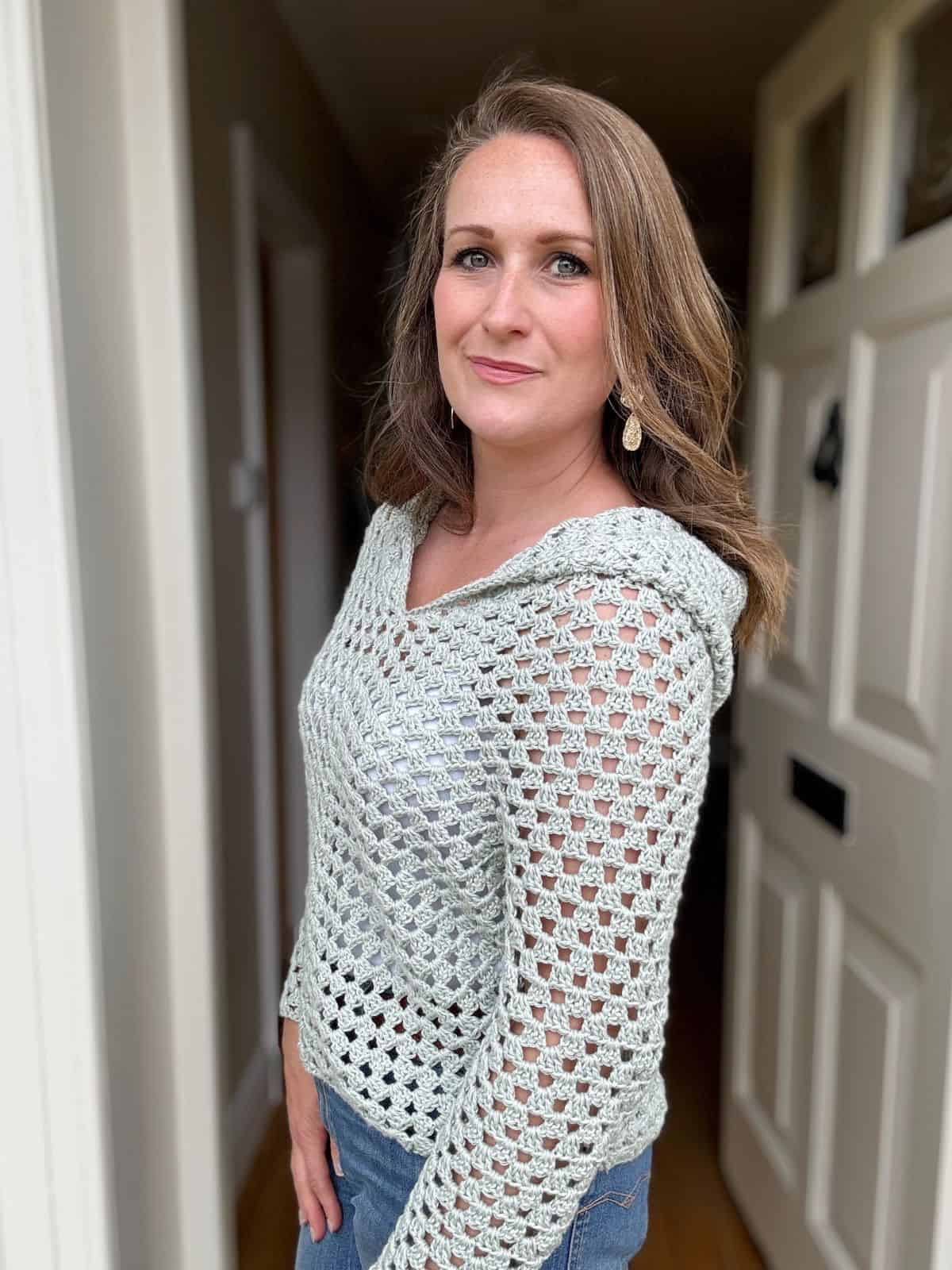 A woman with long brown hair is standing near an open door, wearing a light gray crocheted sweater and blue jeans. She is looking directly at the camera.