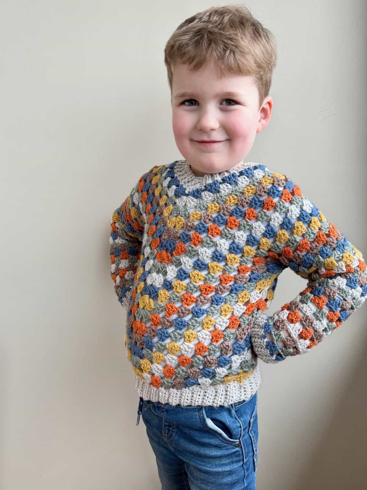 A young child stands against a beige wall, wearing a colorful crocheted sweater and blue jeans, with hands on hips and a slight smile.