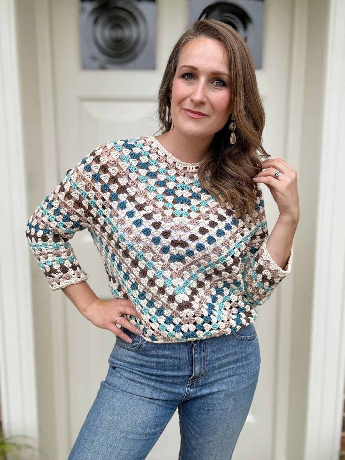 A woman stands in front of a door, wearing a multicolored, crocheted sweater with long sleeves and blue jeans.