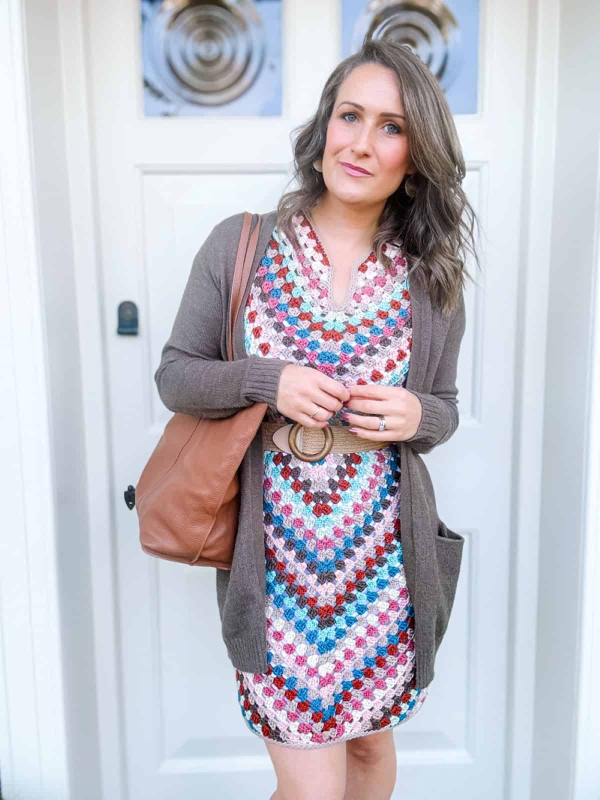 A woman stands in front of a white door, wearing a multicolored summer crochet dress pattern, a brown cardigan, and carrying a brown shoulder bag.