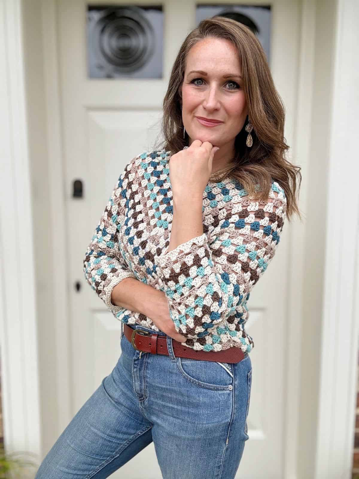 A woman with long hair standing in front of a white door, wearing a stripe crochet sweater, jeans, and a belt, with one hand resting on her chin and the other on her hip.