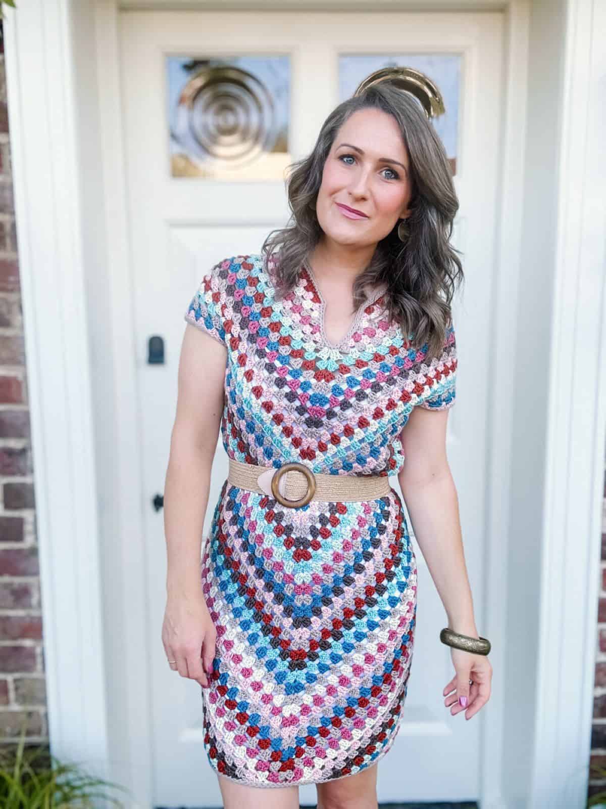 A woman stands in front of a door, wearing a colorful, patterned crochet dress with a beige belt and bracelet.
