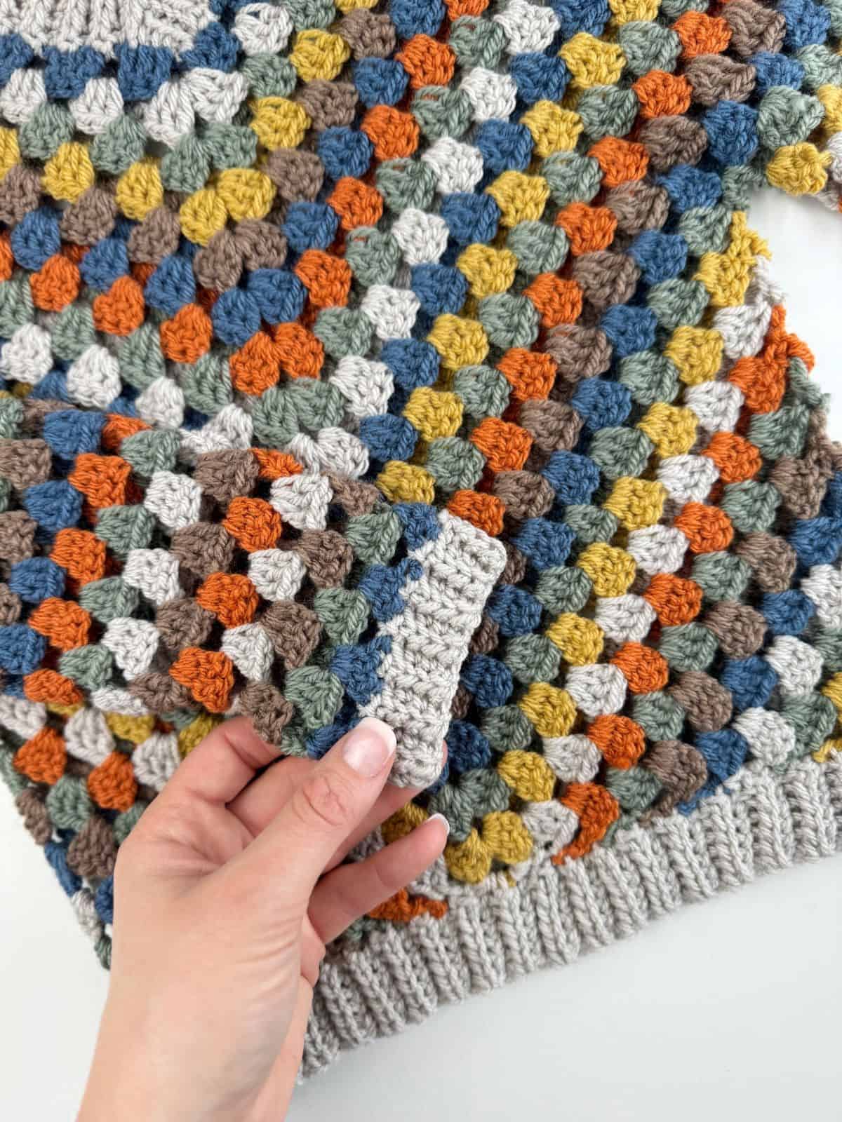 A hand holding a multi-colored crochet piece with a matching multi-colored crochet garment in the background.