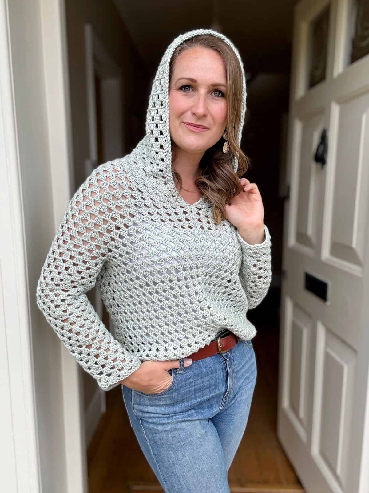 A person stands in front of an open door, wearing a light gray crocheted hoodie and blue jeans, with one hand touching the hood and the other in their pocket. The intricate crochet hoodie pattern adds a touch of handmade charm.