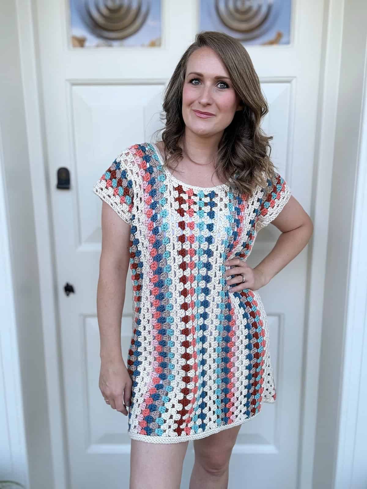 A person stands in front of a white door, wearing a multicolored crochet dress, with one hand on their hip, looking at the camera.