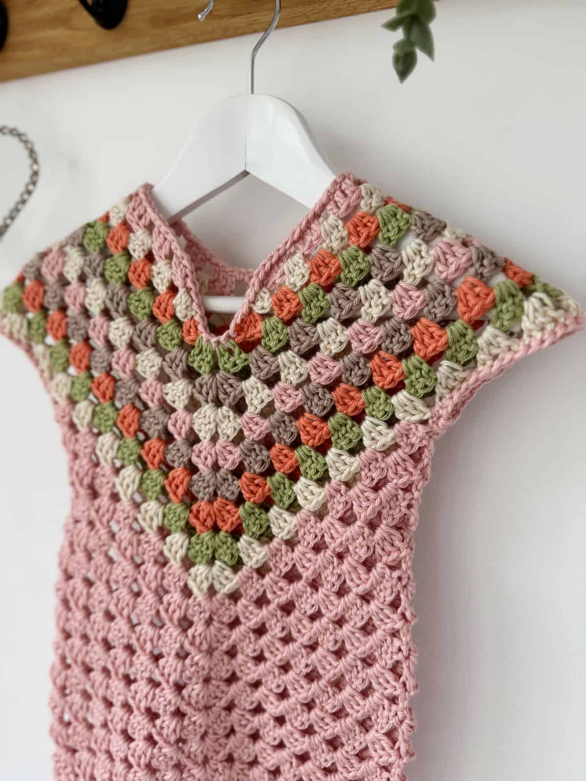 A crocheted dress with a colorful granny square design in shades of pink, green, orange, and beige hangs on a white hanger against a white background, showcasing a charming crochet dress pattern for baby enthusiasts.