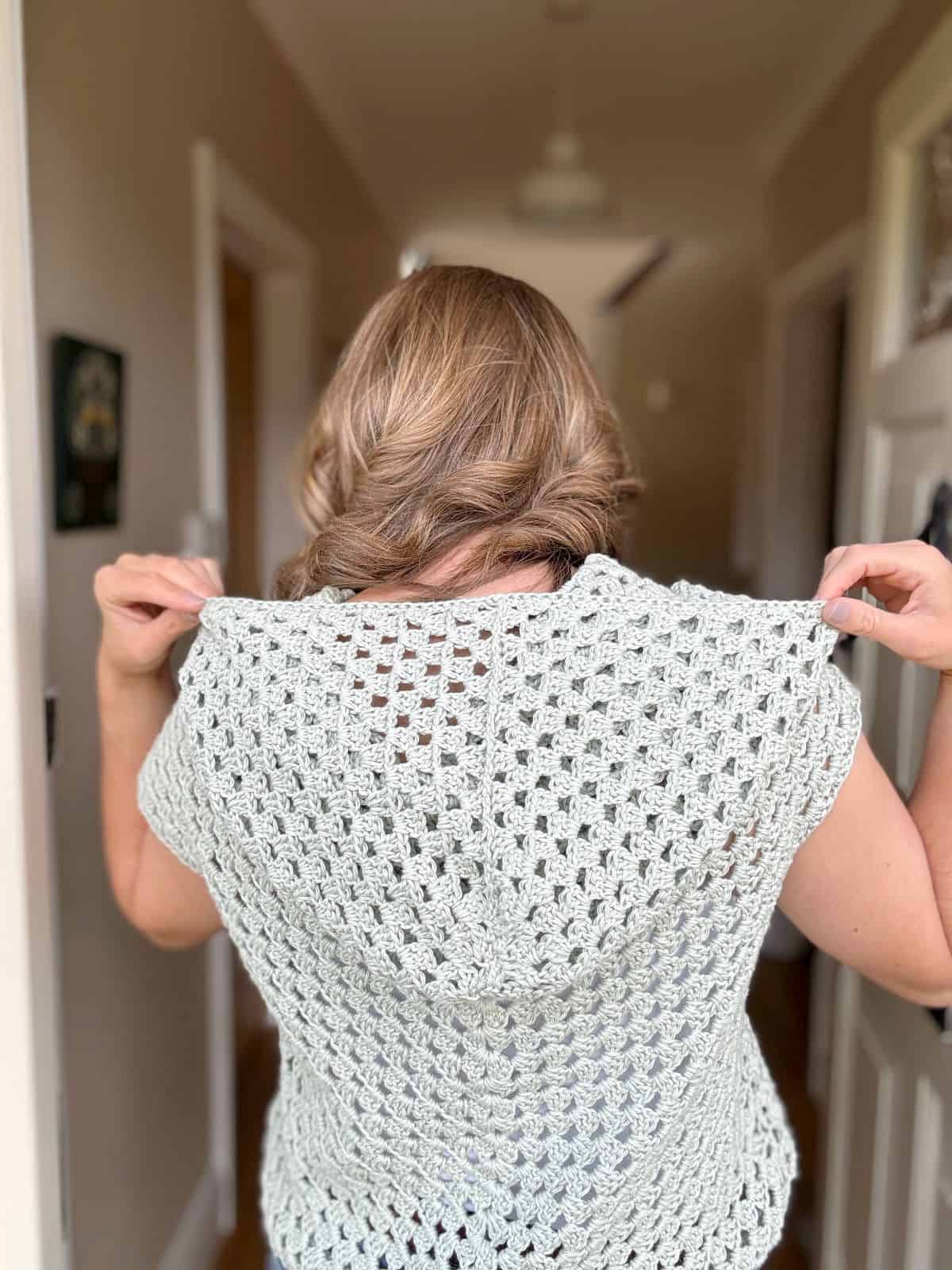 A person with light brown hair stands indoors, facing away, showcasing a light green crocheted garment over their shoulders, hinting at a stylish crochet hoodie pattern.