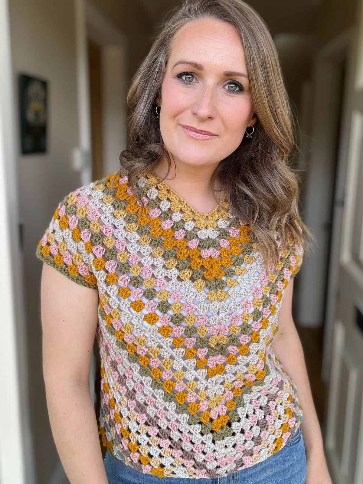 A woman wearing a colorful, patterned crochet top stands indoors, looking at the camera with a neutral expression.