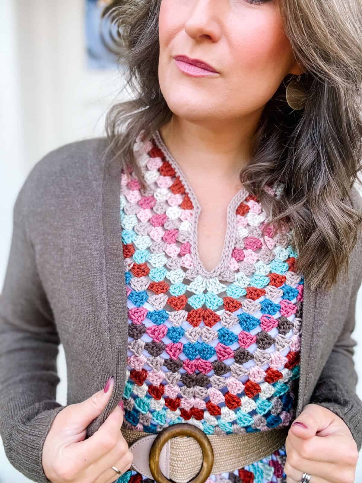 A person wearing a multicolored crocheted dress with a summer crochet dress pattern, a brown cardigan, and a brown belt with a large circular buckle. Only head and torso are visible.