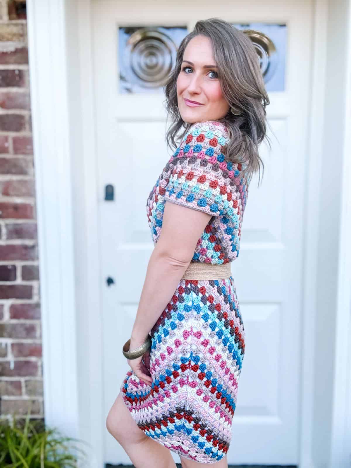 A woman stands in front of a white door and brick wall, wearing a summer crochet dress pattern with a beige belt. She has shoulder-length hair and is looking over her shoulder at the camera.