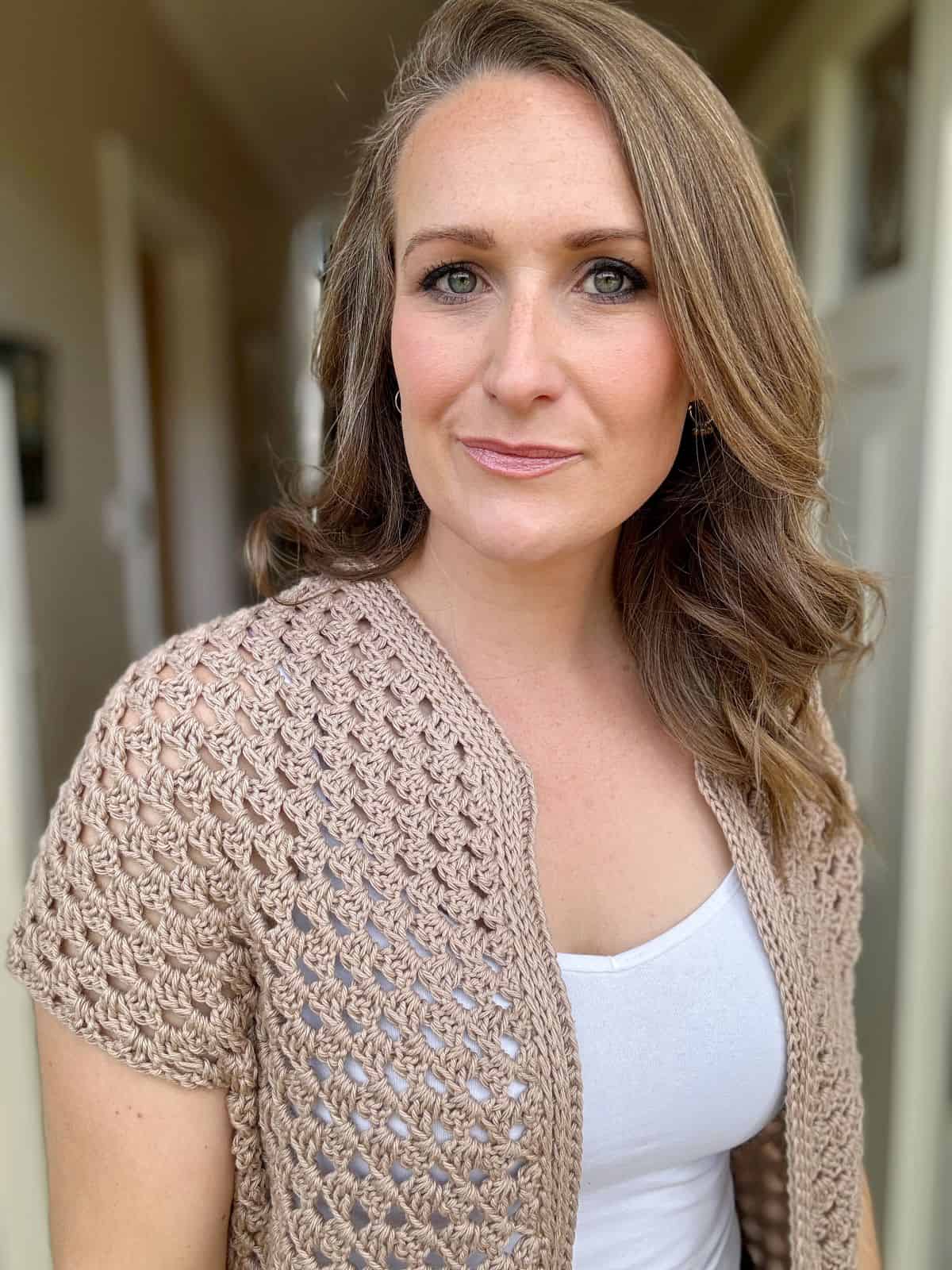 A person with light brown, wavy hair wearing a white top under a beige granny stitch cardigan stands in a hallway, looking directly at the camera.