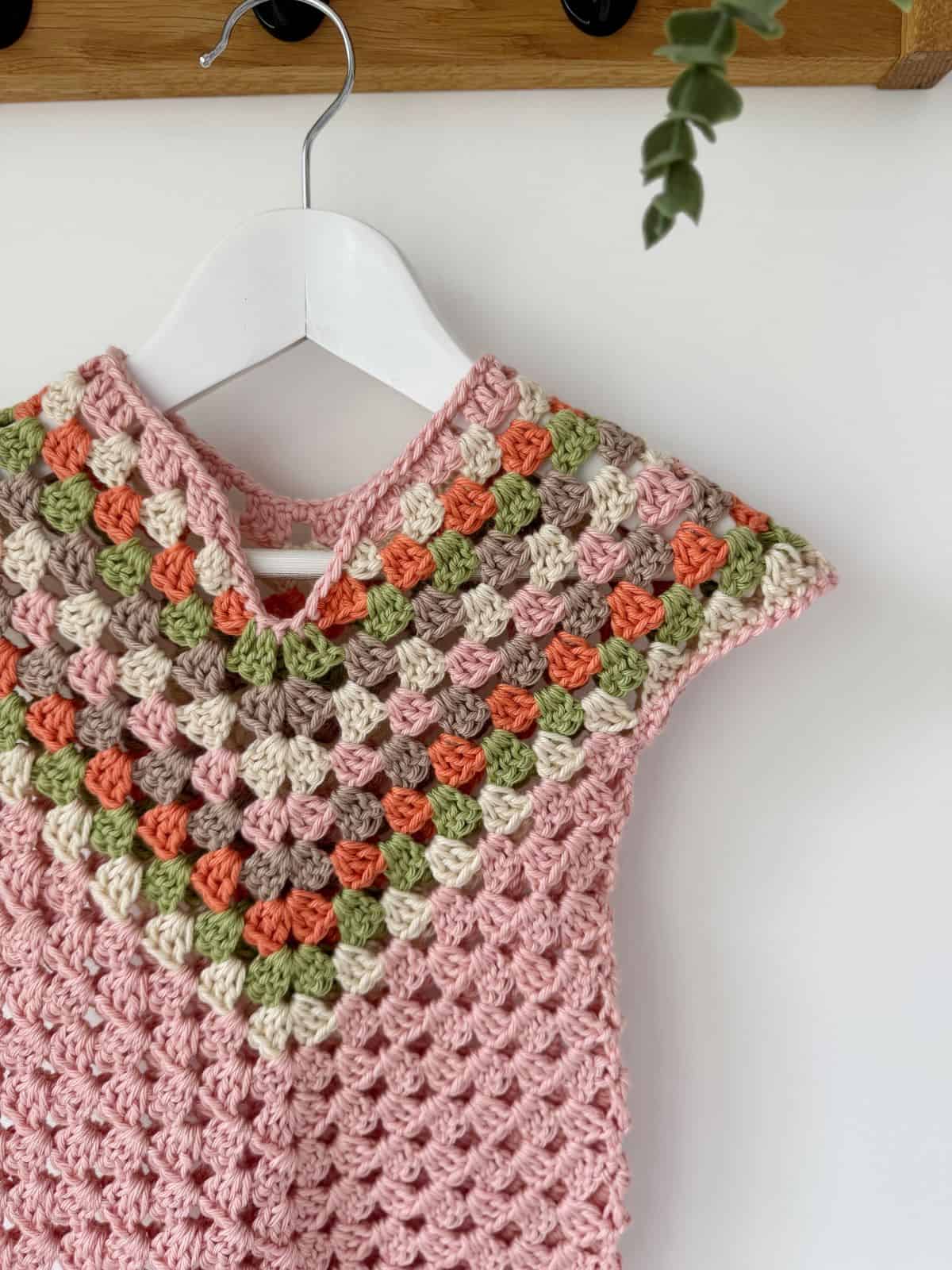 A multicolored crocheted dress with a diamond-shaped pattern in pink, green, orange, and brown, resembling a cozy crochet dress pattern for baby, hangs gracefully on a white hanger.