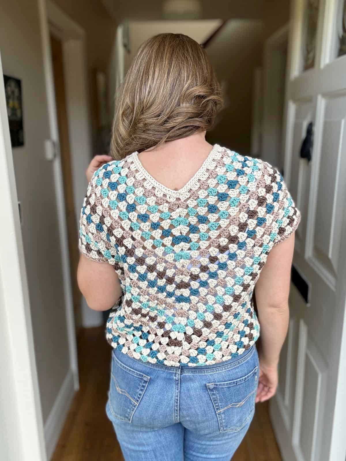 Person with wavy, blonde hair facing away, wearing a multicolored crochet top and blue jeans. They are standing in a hallway with light-colored walls and doors on either side.