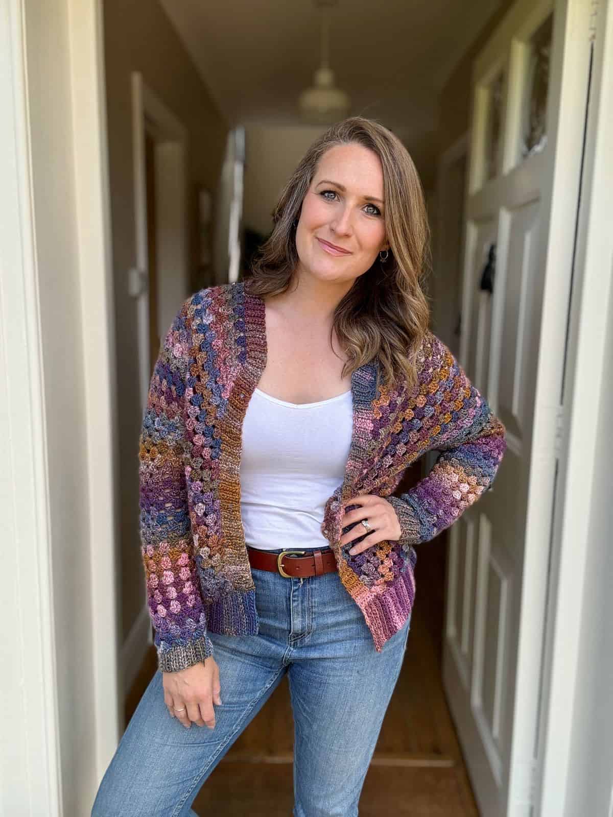 A woman stands in a hallway wearing a multicolored, granny stitch cardigan over a white tank top and jeans, with one hand in her pocket. She has wavy hair and gently smiles at the camera.