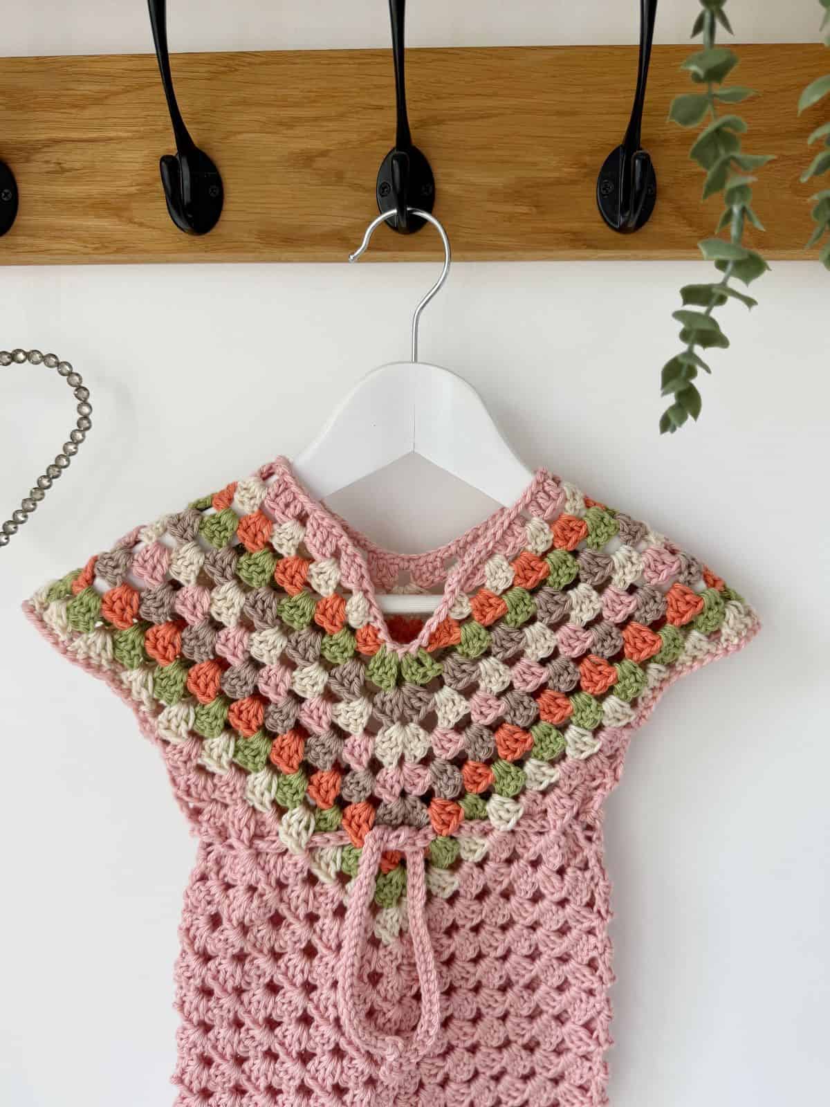 A crochet dress pattern for baby showcases a multicolored design, hanging gracefully on a white hanger placed on a hook rack against a pristine white wall.