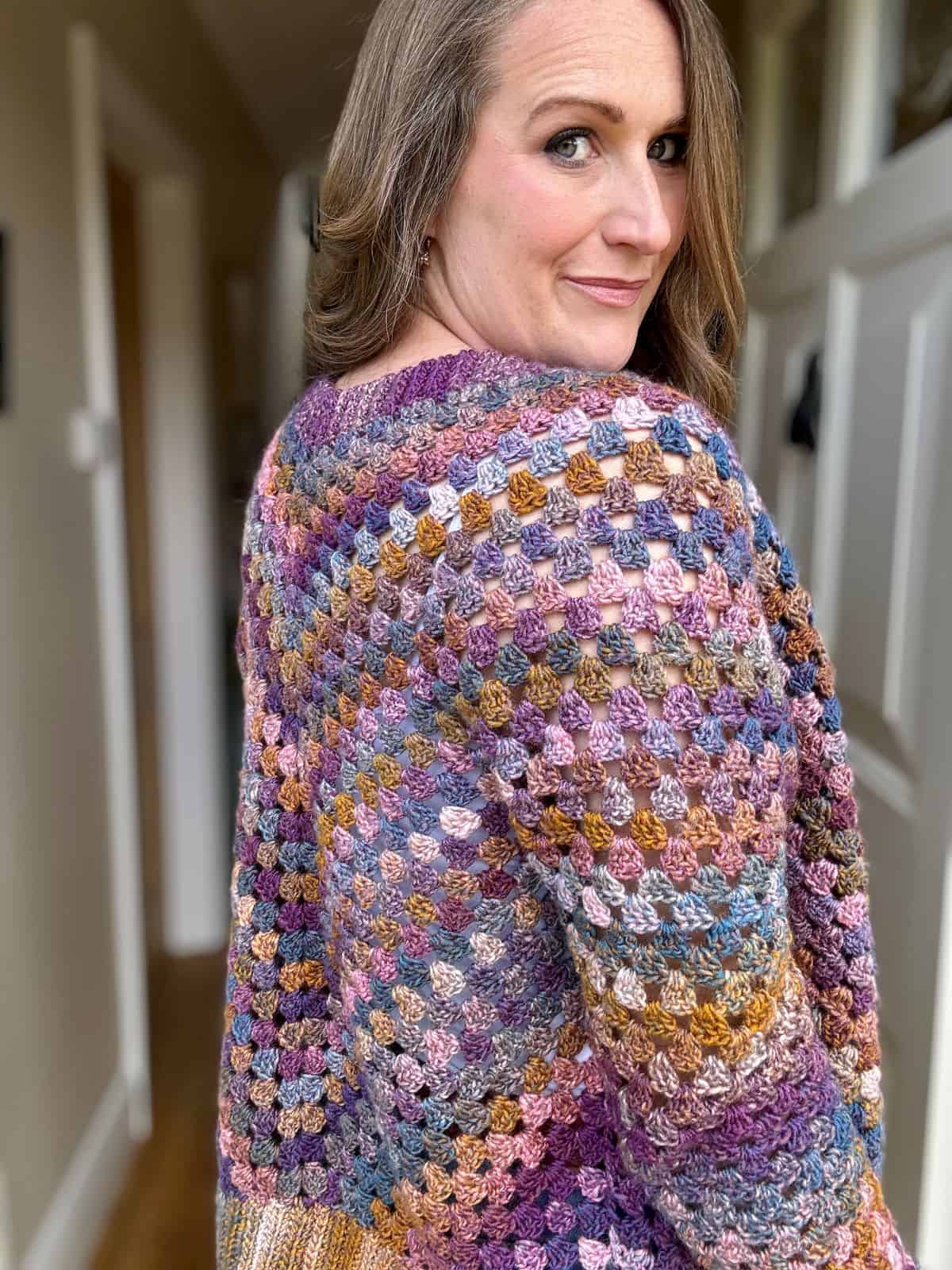 A person with long hair wearing a colorful, granny stitch cardigan stands indoors, looking over their shoulder.