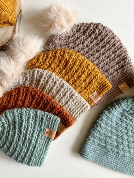 Five crocheted beanies in various colors, including blue, orange, mustard, beige, and brown with a pom-pom, are laid out on a white surface.