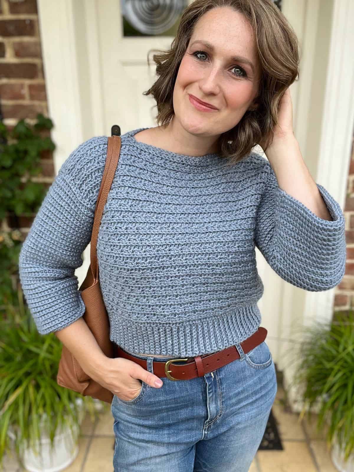 A woman with short brown hair, wearing a light blue V-neck crochet sweater pattern, blue jeans, and a brown belt, stands in front of a door, holding a brown shoulder bag. She has a slight smile and touches her hair.