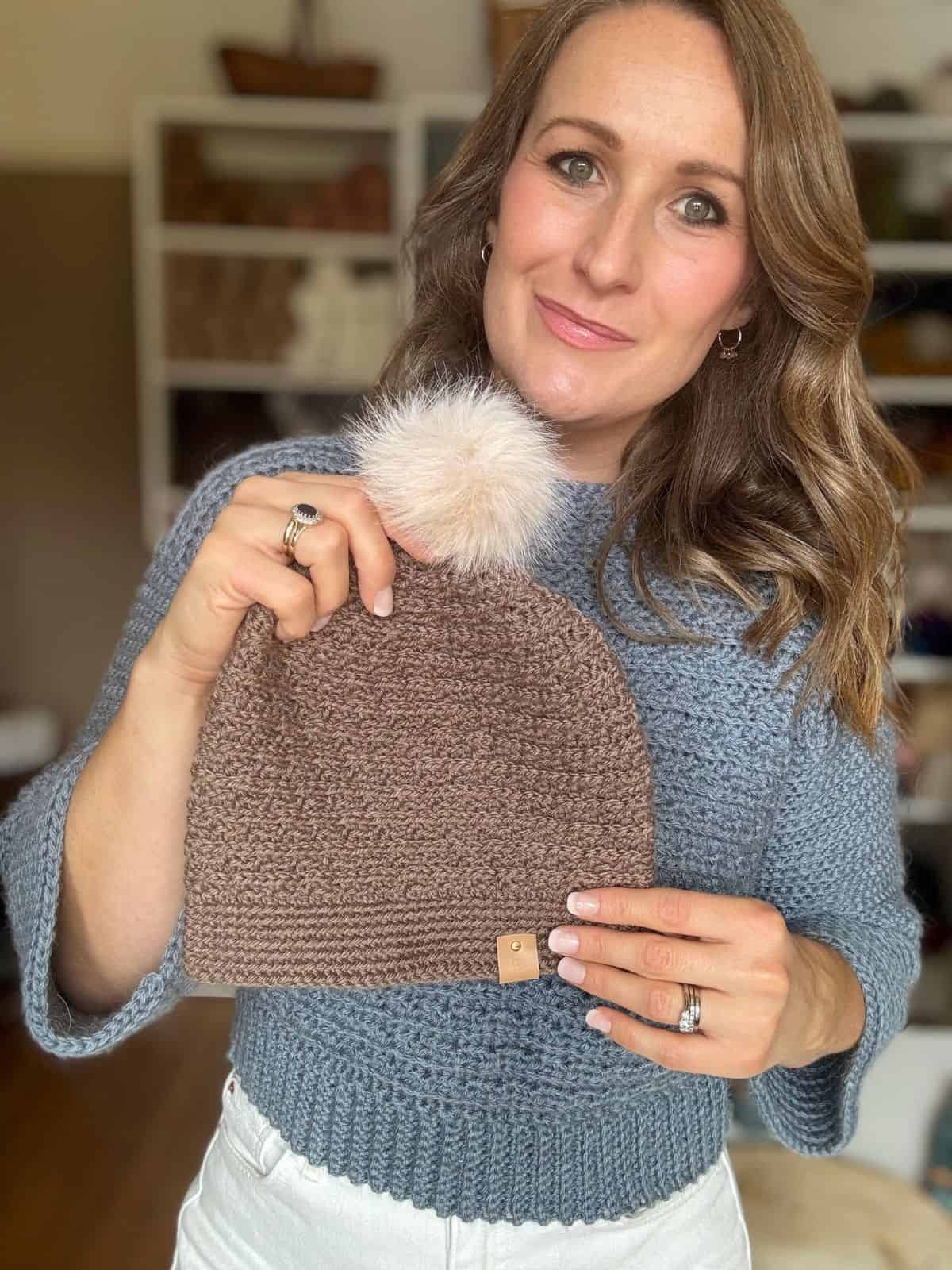 A woman in a blue sweater holds up a brown crochet beanie with a pom-pom, showcasing a boys crochet hat pattern, in a room with shelves in the background.