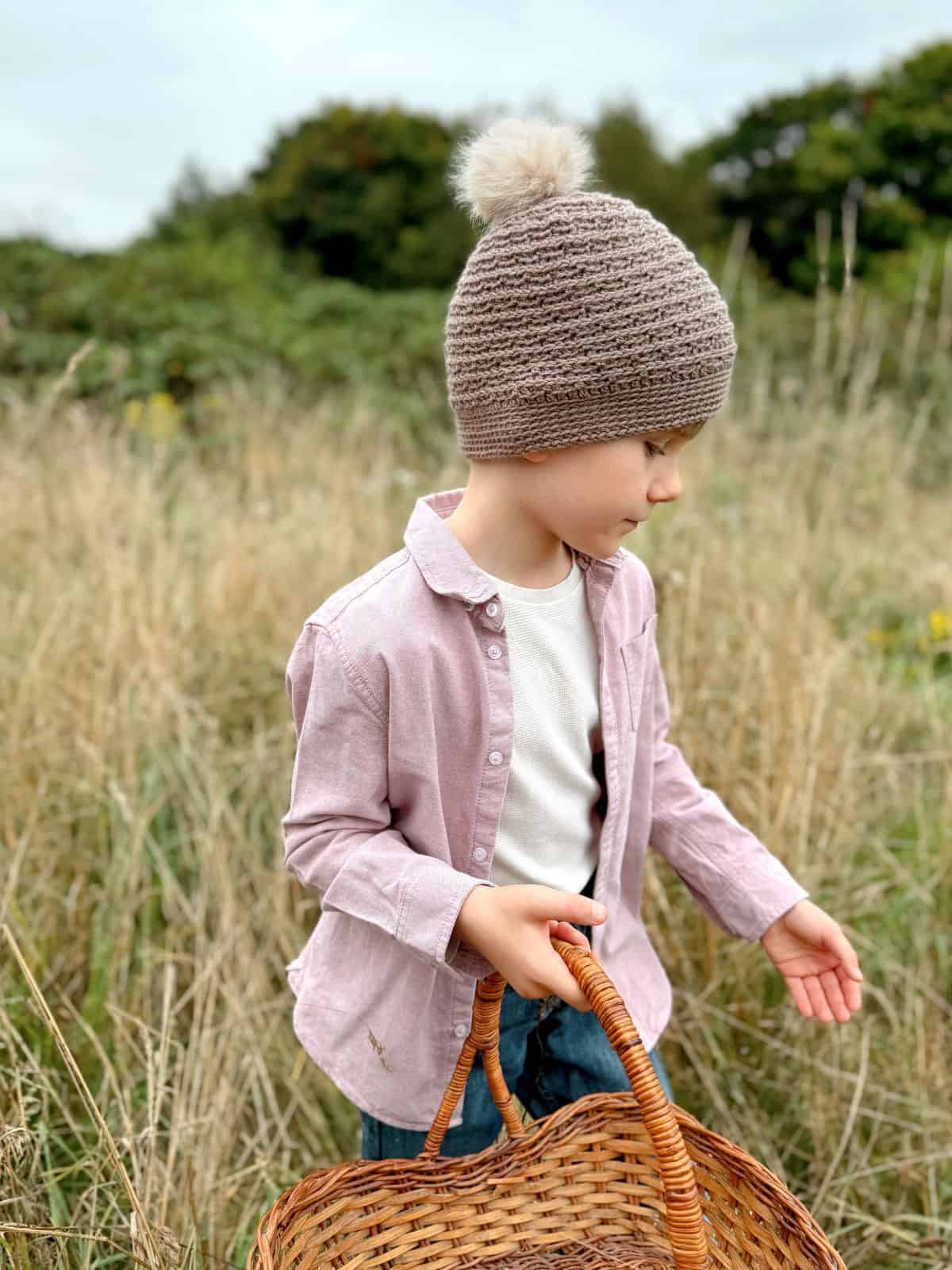 A child wearing a boys crochet hat pattern and a pink shirt holds an empty wicker basket while standing in a grassy field.