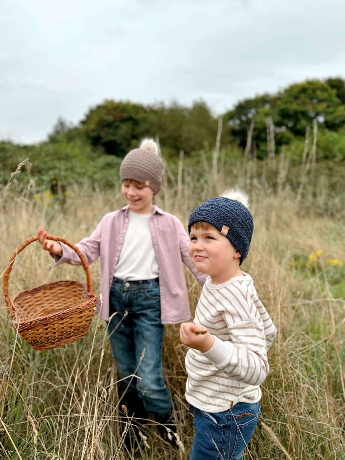 Two young children, both wearing crochet hats, are standing in a grassy field. The older one holds a wicker basket and they both look content. The scene might have been inspired by a boys crochet hat pattern. Trees and cloudy sky are in the background.