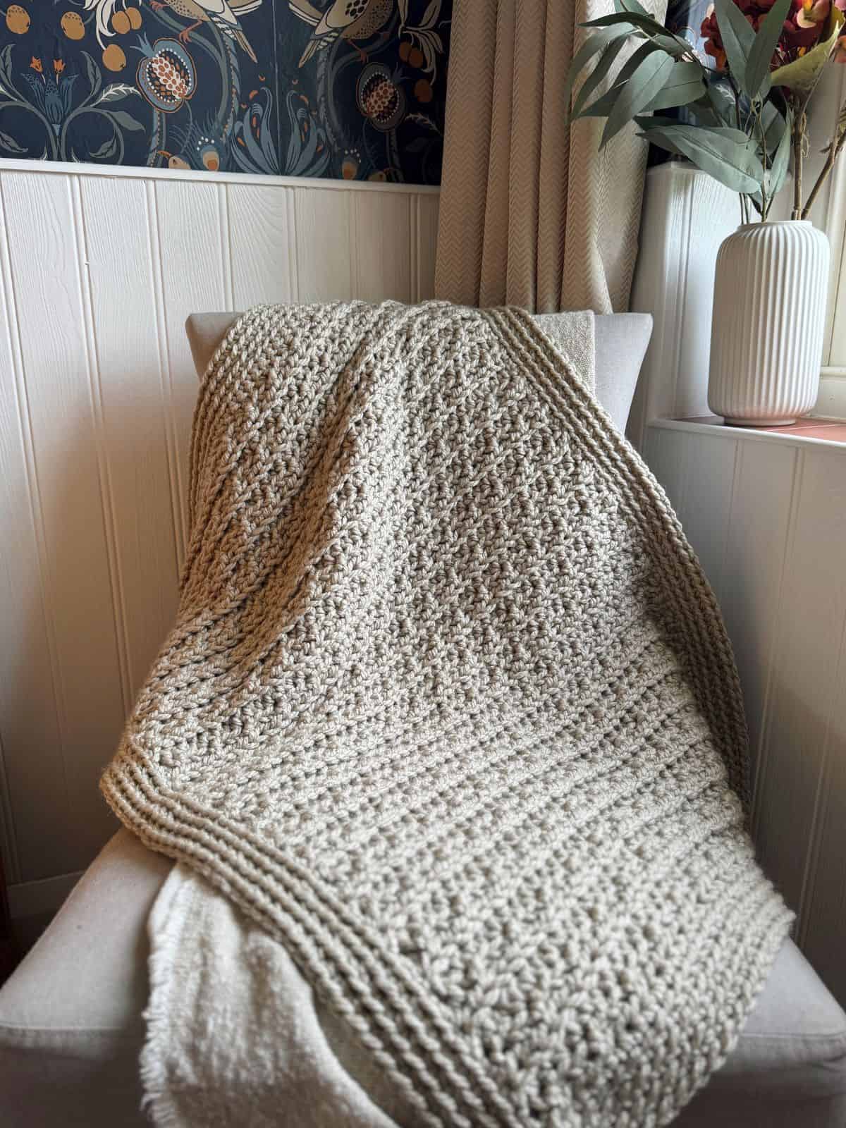 A chunky crochet blanket pattern, featuring a textured beige design, is draped over a light-colored chair in a room with floral-patterned wallpaper and a white vase with greenery on the windowsill.