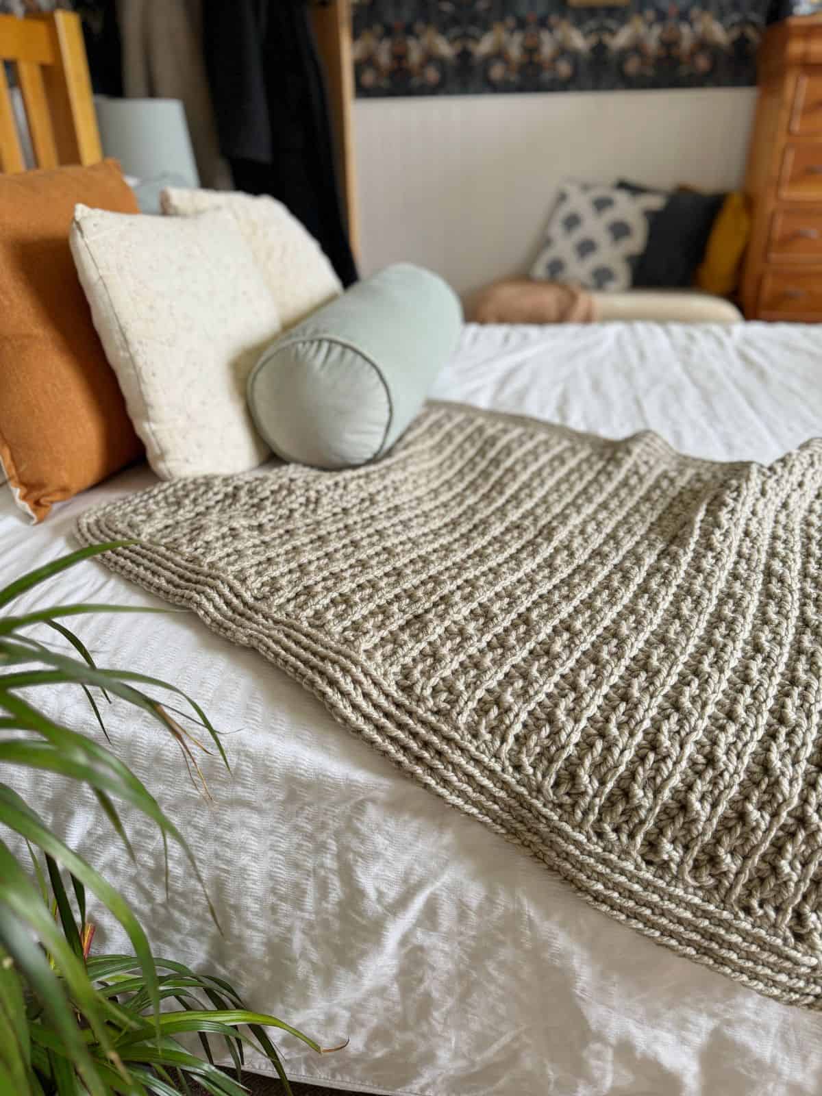 A neatly made bed with a chunky crochet blanket pattern draped over the foot, flanked by various pillows, including a cylindrical bolster and a lush green plant nearby.