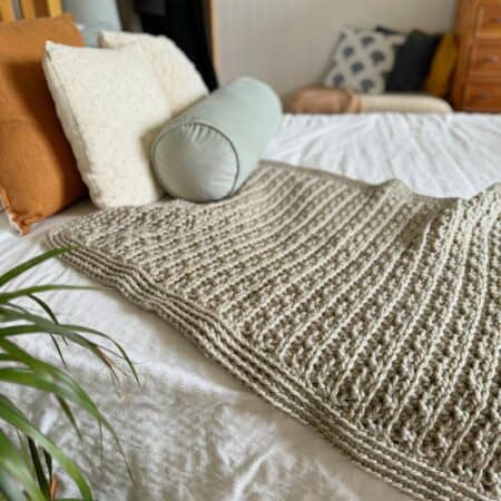 A neatly made bed with a textured crochet blanket, assorted pillows, and a plant in the foreground.