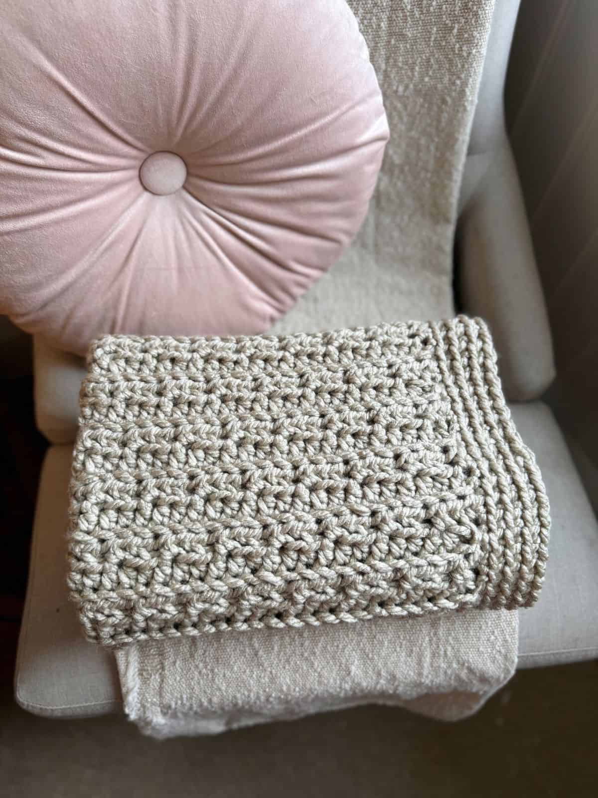 A cream-colored blanket, made from a chunky crochet blanket pattern, is neatly folded on a chair with a pink round cushion.