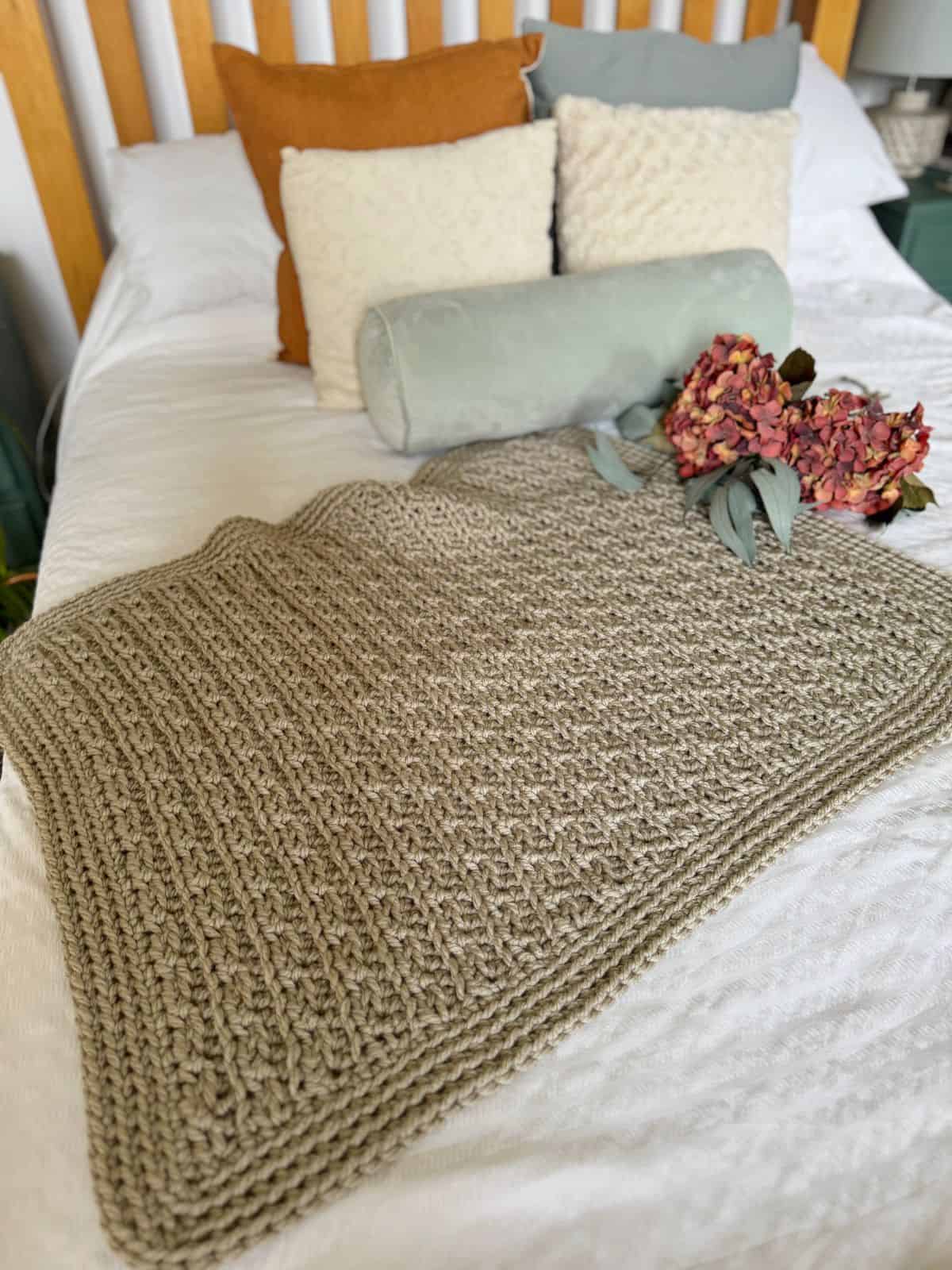 A beige chunky crochet blanket patterned with care is laid out on a neatly made bed with white sheets, surrounded by pillows in various colors and a bouquet of dried flowers.