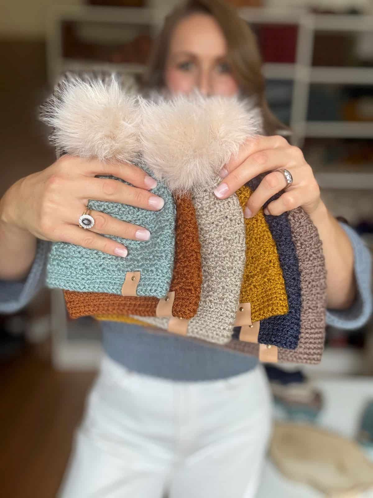 Person holding a collection of six crocheted hats in various colors with fluffy pom-poms, showcasing an array of boys crochet hat patterns.