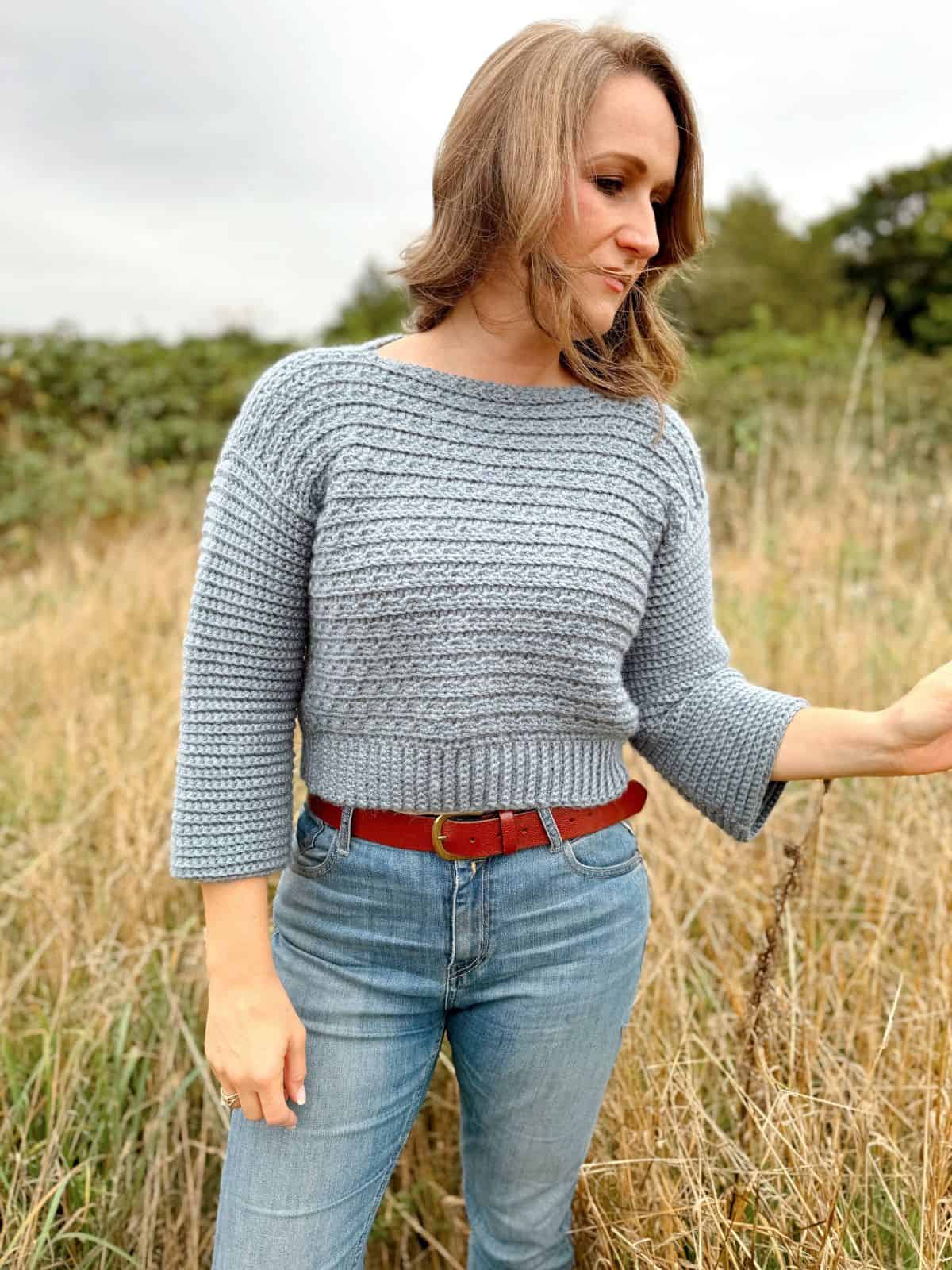 A woman wearing a grey V-neck crochet sweater and jeans stands in a grassy field, looking down and to the side.