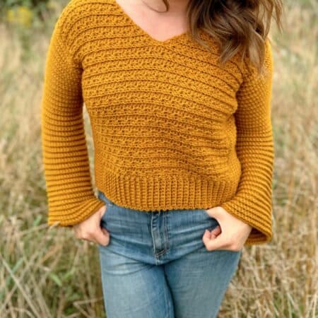 Person wearing a mustard yellow V-neck crocheted sweater and blue jeans, standing in a grassy field.