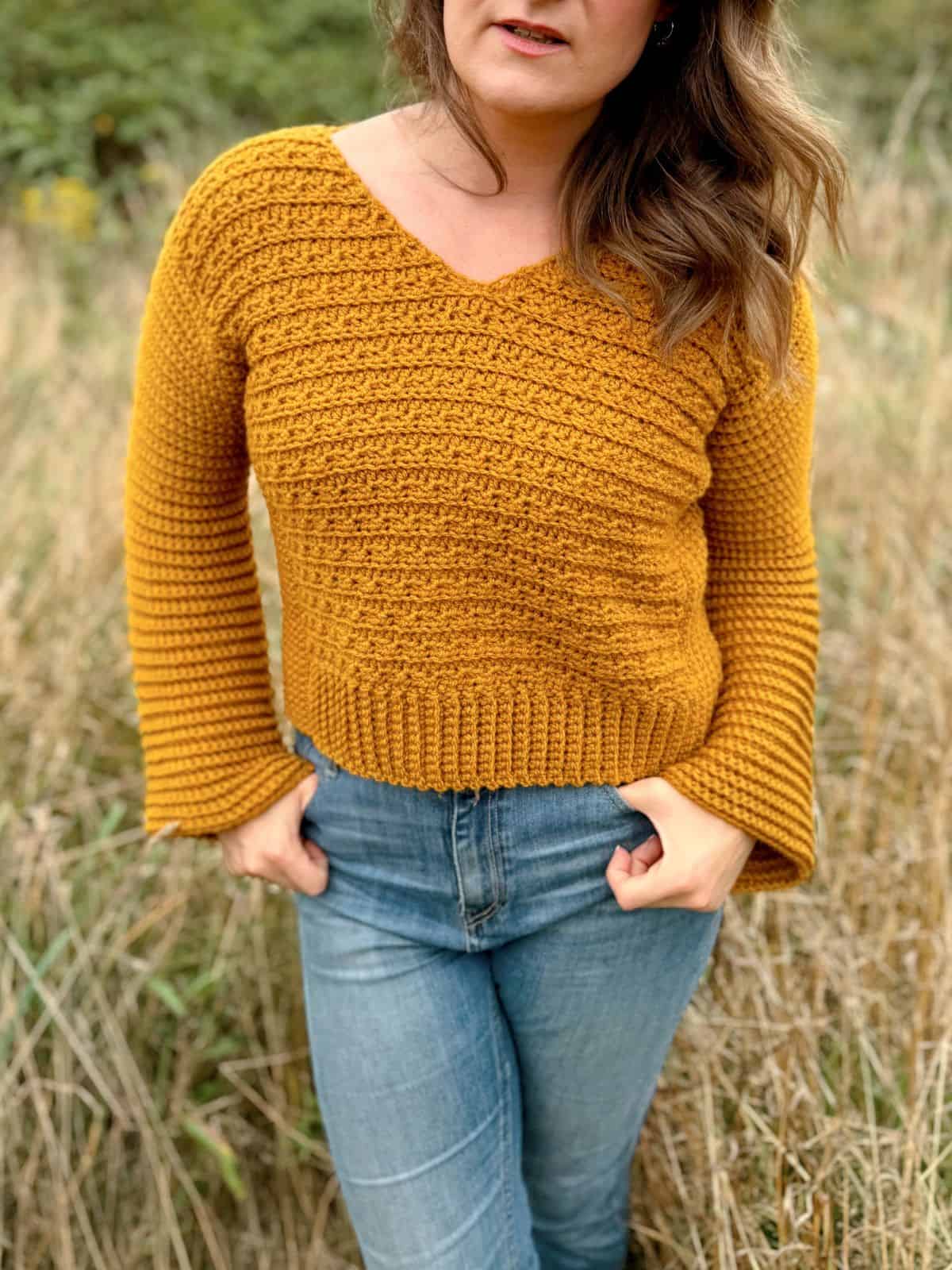 Person wearing a mustard yellow V-neck crocheted sweater and blue jeans, standing in a grassy field.