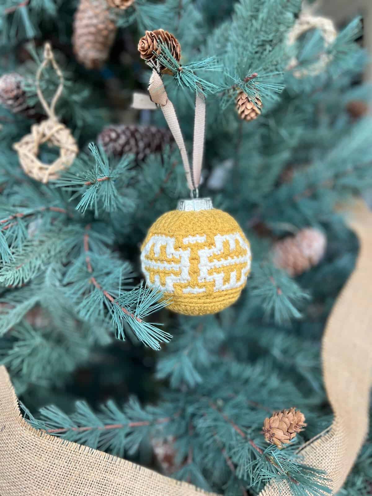 A yellow and white patterned crochet bauble, resembling an easy mosaic crochet design, hangs on a Christmas tree decorated with pinecones and other natural elements.