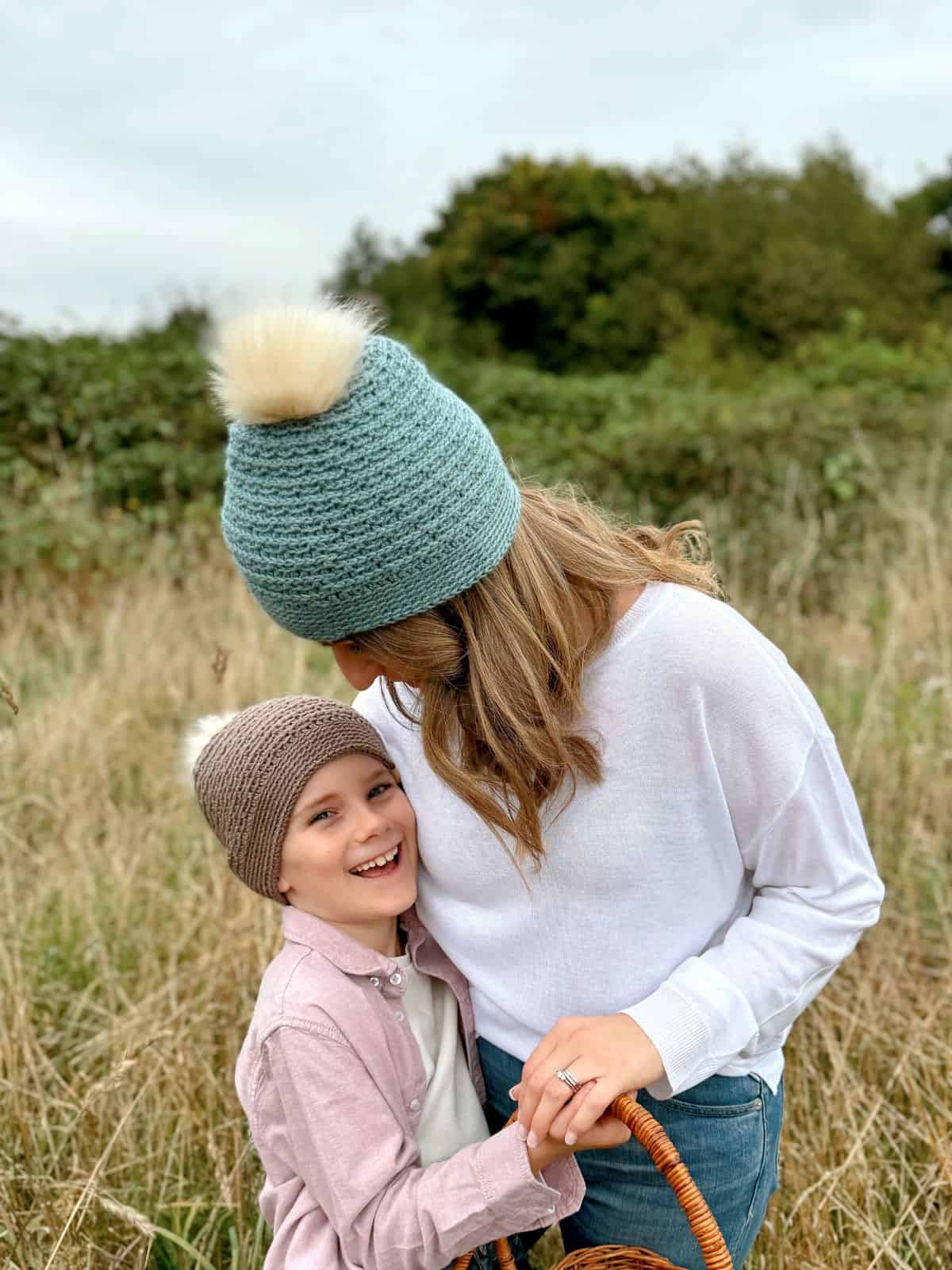 An adult and a child, both wearing boys' crochet hats with pom-poms, stand together in a grassy field. The adult holds a basket and leans down, while the child smiles at the camera.