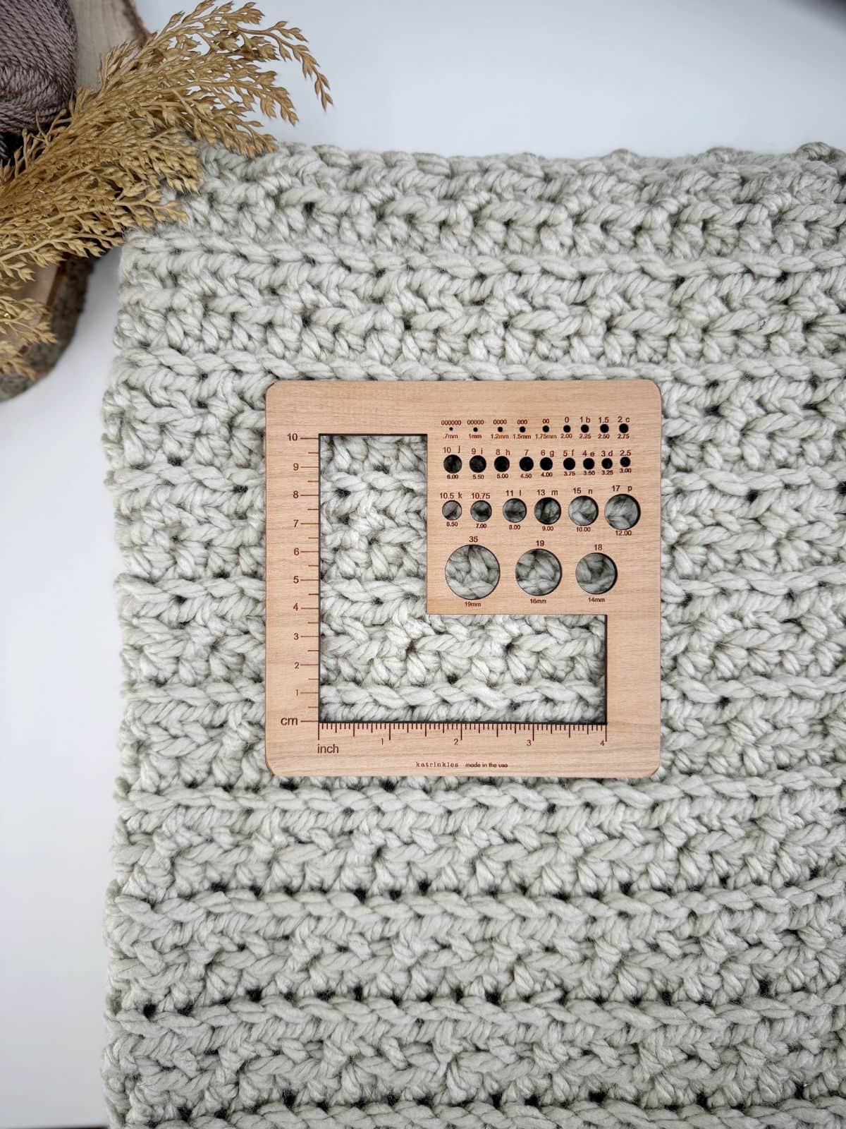 A wooden crochet gauge tool rests on a chunky, gray crochet fabric. Perfect for your chunky crochet blanket pattern, the tool includes various needle-sized holes, measurements in inches and centimeters, and convenient conversion charts.