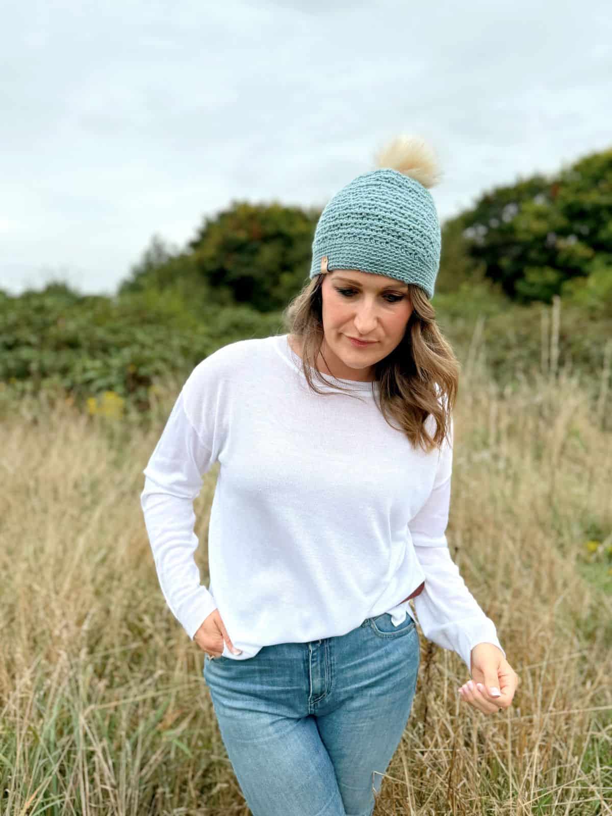 A woman wearing a light blue crochet beanie and white long-sleeve shirt stands in a grassy field with trees in the background, reminiscent of a boys' crochet hat pattern.