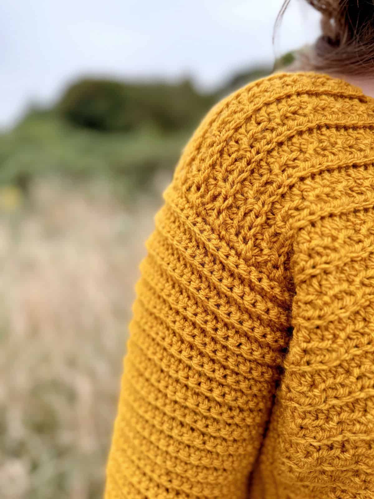 Close-up of a person wearing a mustard yellow, V-neck crochet sweater with a nature background. The image focuses on the crochet pattern and texture of the sweater.