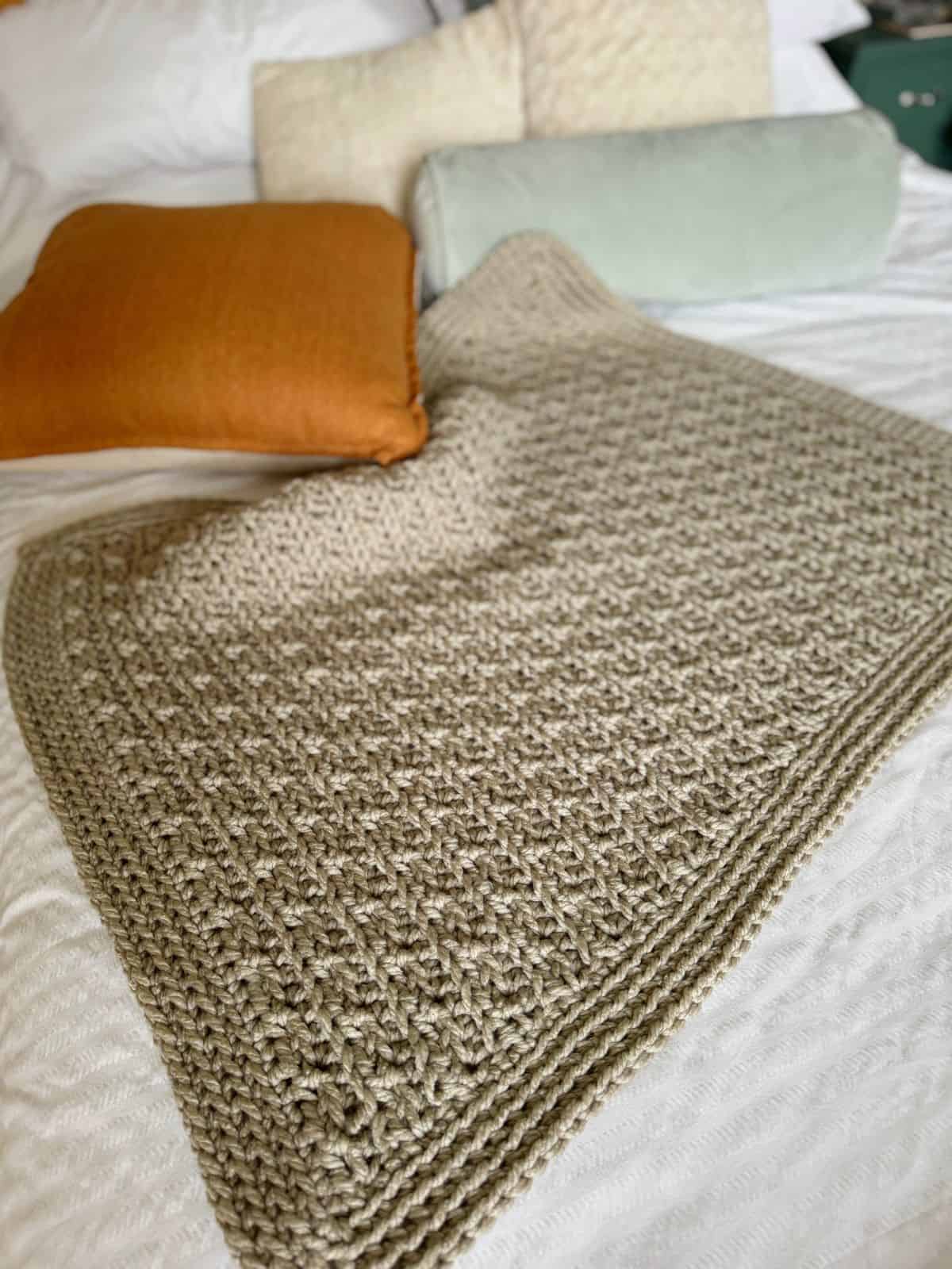 Chunky crochet blanket pattern draped over a bed with three pillows: one orange, one light green, and one off-white. The bed has white sheets.