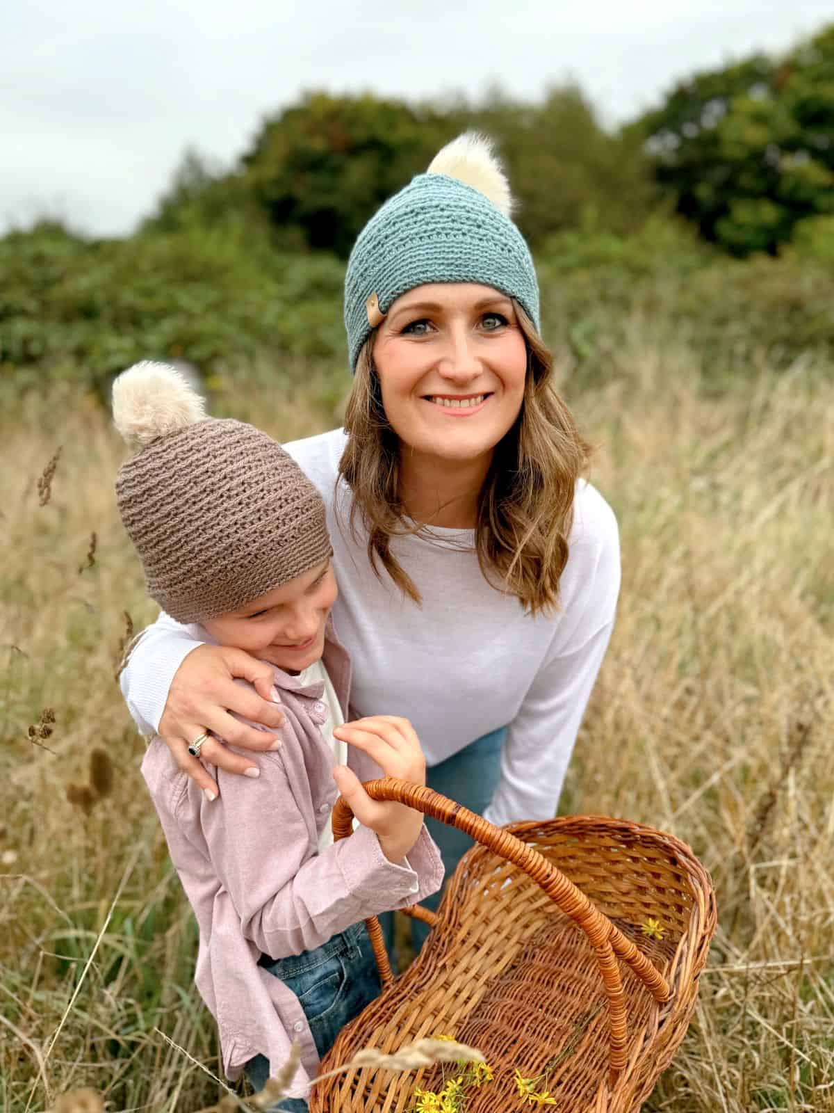 A woman and a child wearing crochet hats stand in a field, holding a wicker basket. The woman smiles while the child, whose hat is made from a boys crochet hat pattern, looks down. The field has tall grasses and trees in the background.