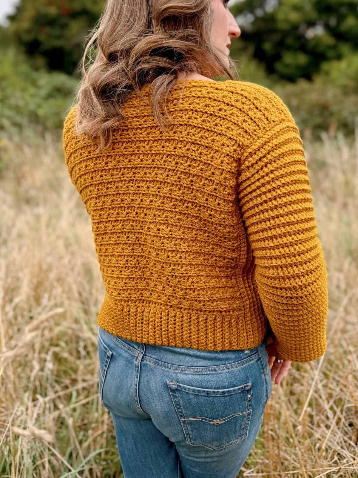 A person wearing a mustard-colored, v-neck crochet sweater and blue jeans stands in a grassy field with trees in the background.