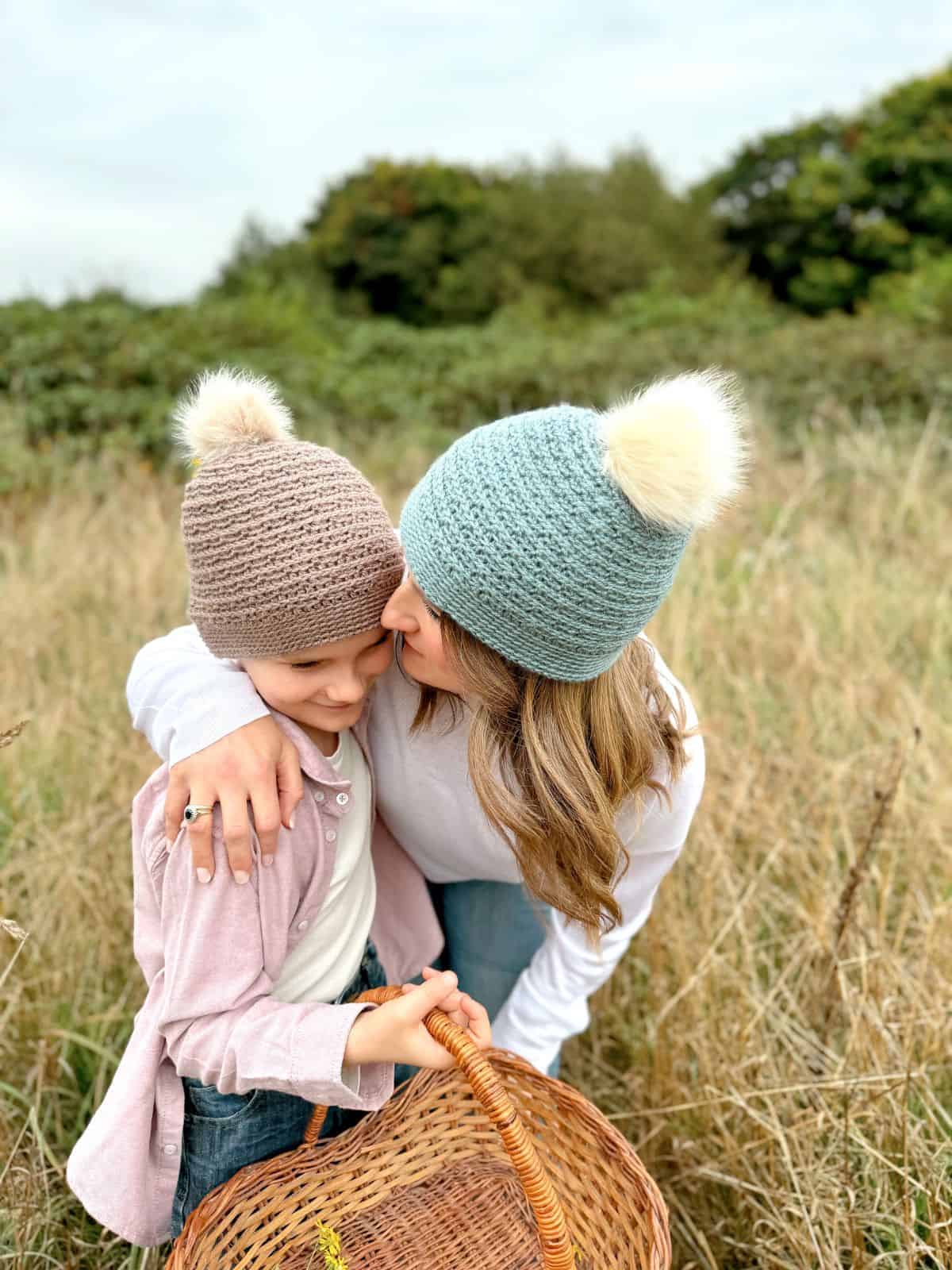 A woman and a child wearing crochet hats with pom-poms stand in a grassy field. The woman kneels, hugging and kissing the child on the cheek, while the child holds a woven basket, showcasing what could be an adorable boys crochet hat pattern.