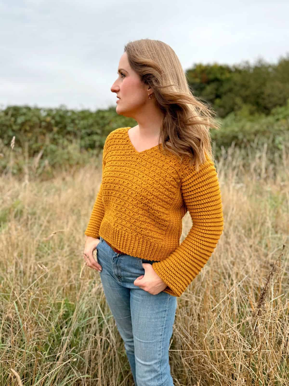 Person standing in a field, wearing a mustard-colored v-neck crochet sweater pattern and blue jeans, with hands in pockets and looking to the left.