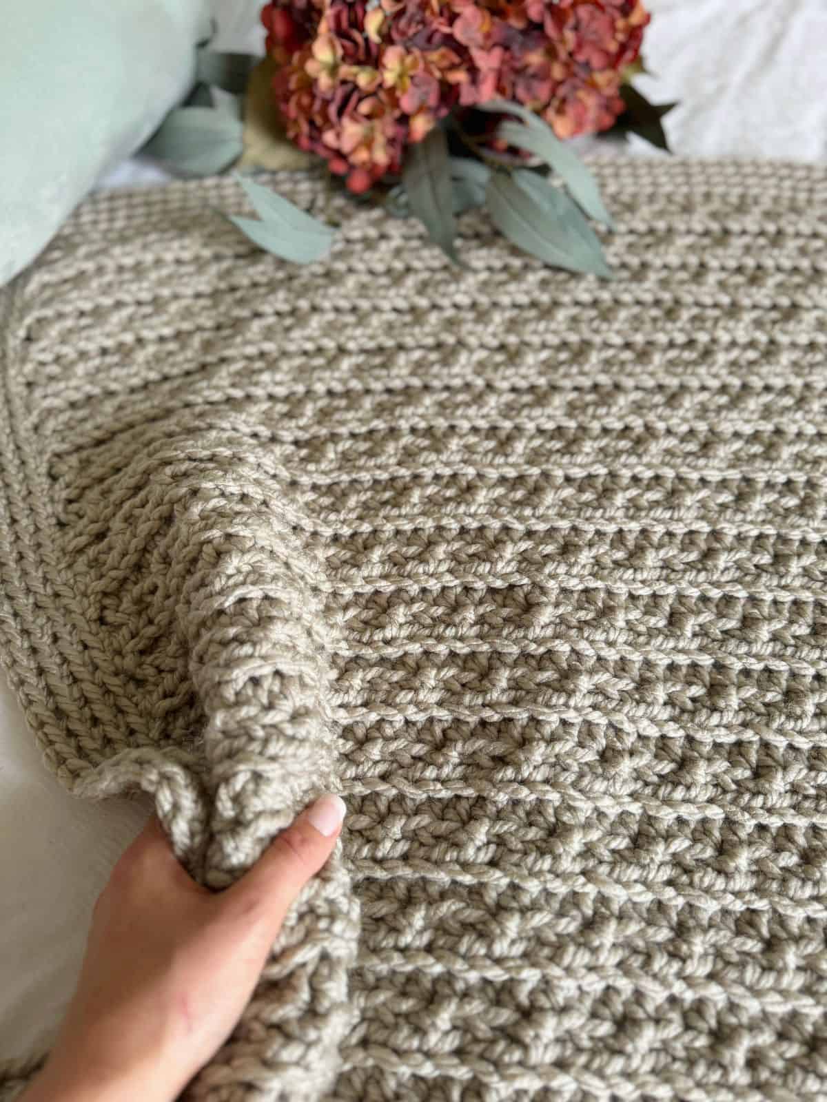 A hand holds up a chunky crochet blanket, its thick beige threads draping over the bed, with a bouquet of dried flowers in the background.