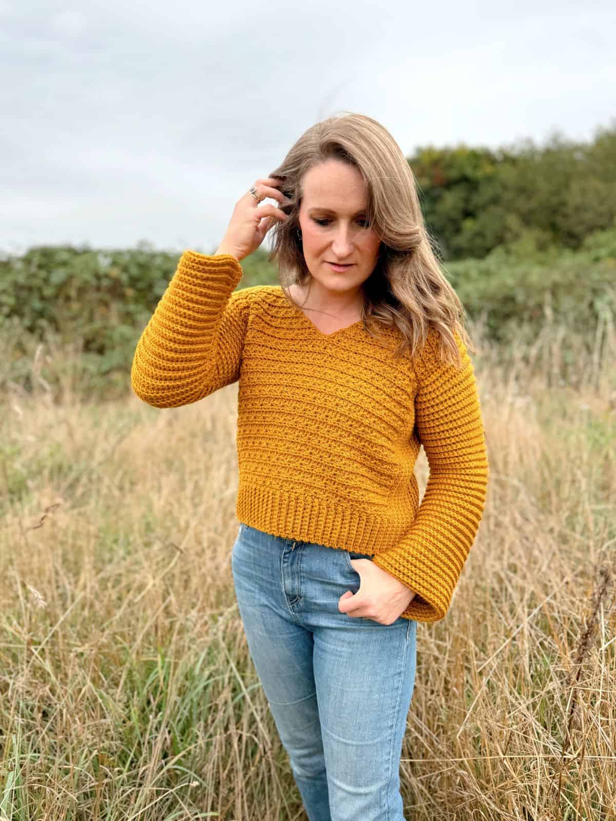 A person with long hair stands in a grassy field, wearing a mustard yellow V-neck crochet sweater pattern and blue jeans, looking down while touching their hair. Trees and shrubs are in the background under a cloudy sky.