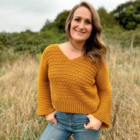 A person stands outside in a grassy area, wearing a mustard-colored v-neck crochet sweater and blue jeans, smiling at the camera.