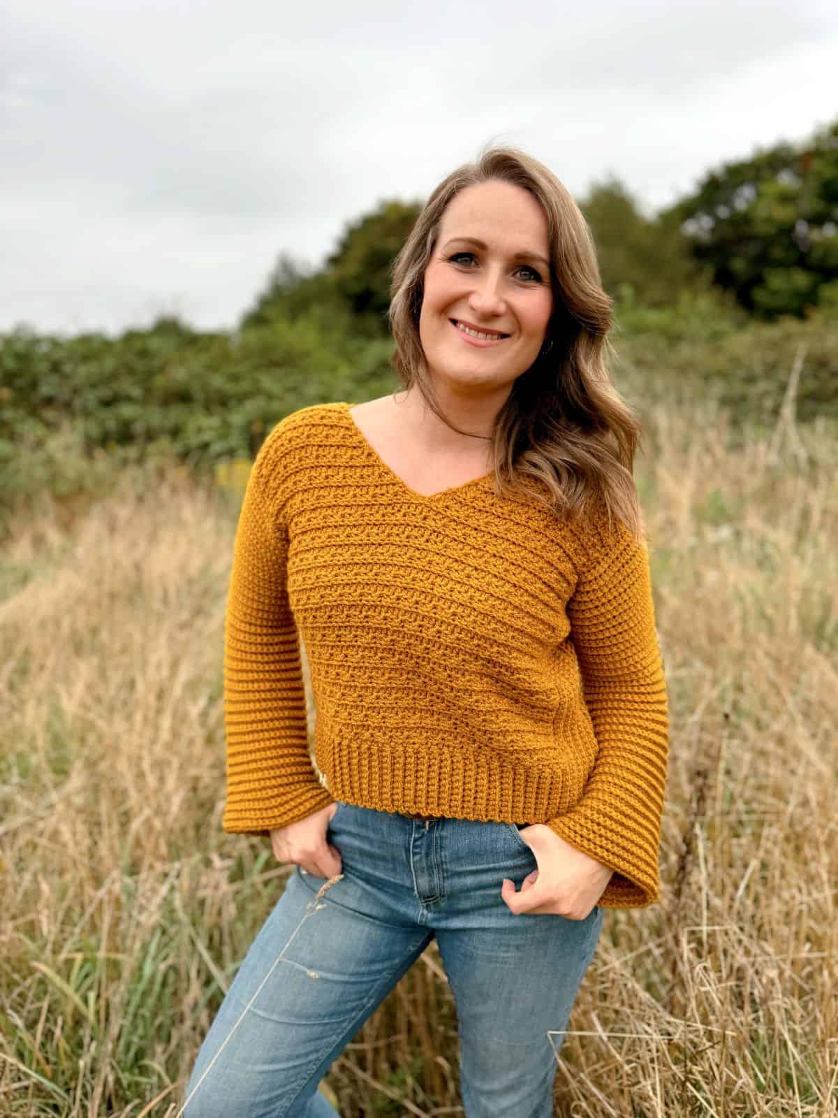 A person stands outside in a grassy area, wearing a mustard-colored v-neck crochet sweater and blue jeans, smiling at the camera.