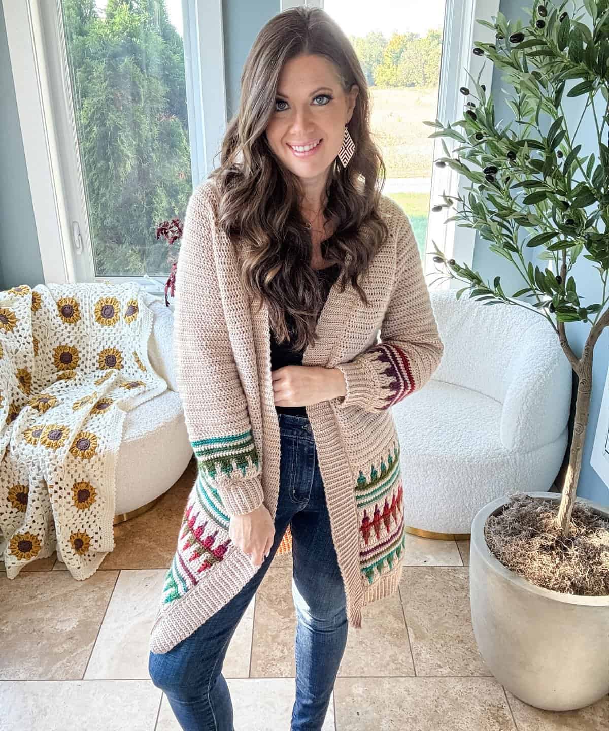 A person with long hair wearing a beige cardigan with colorful patterns stands indoors near a potted plant and a white chair.